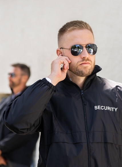 Agent de sécurité en veste noire marquée SECURITY, lunettes de soleil, écoute son oreillette. Un autre agent derrière.