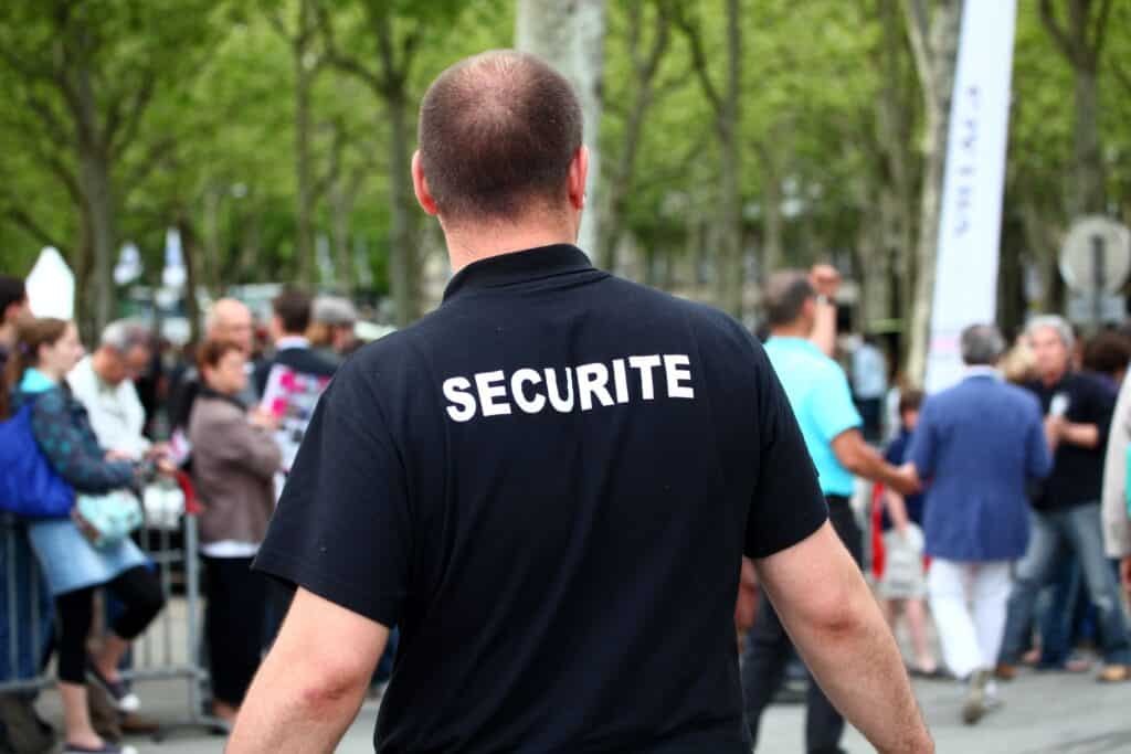 Agent SECURITE : Surveillance de foule en événement extérieur. Personne vue de dos avec polo noir "SECURITE", parmi une foule en extérieur avec arbres et barrières.