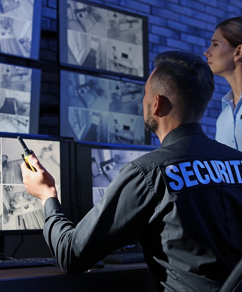 Agent de sécurité avec talkie-walkie et une femme surveillent plusieurs écrans de caméras de surveillance. Inscription SECURITY au dos.