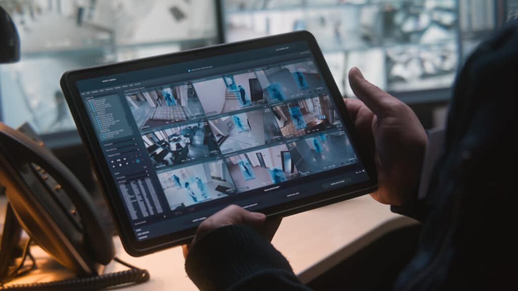 Contrôle surveillance : une personne sur tablette regarde des flux de caméras avec détection IA. Fonds d'écrans : 'SURVEILLANCE SYSTEM PROGRAM'.